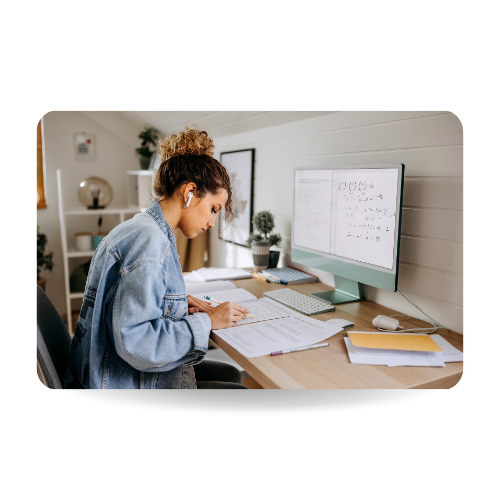 There is a girl in a desk studying in front of the computer and writing in a notebook